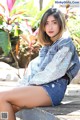 A woman sitting on a wooden bench in a denim jacket and shorts.