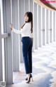 A woman in a white shirt and blue pants leaning against a wall.